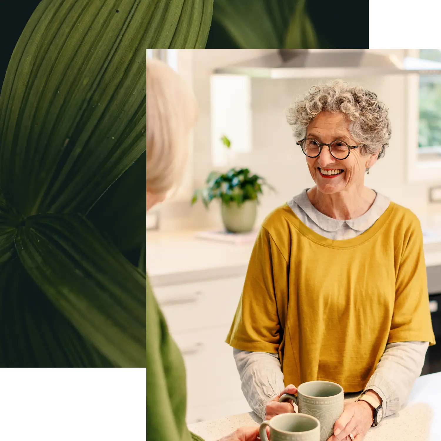 A woman with curly grey hair and glasses smiling while talking to her friend inside an Olive Estate home, holding a mug.