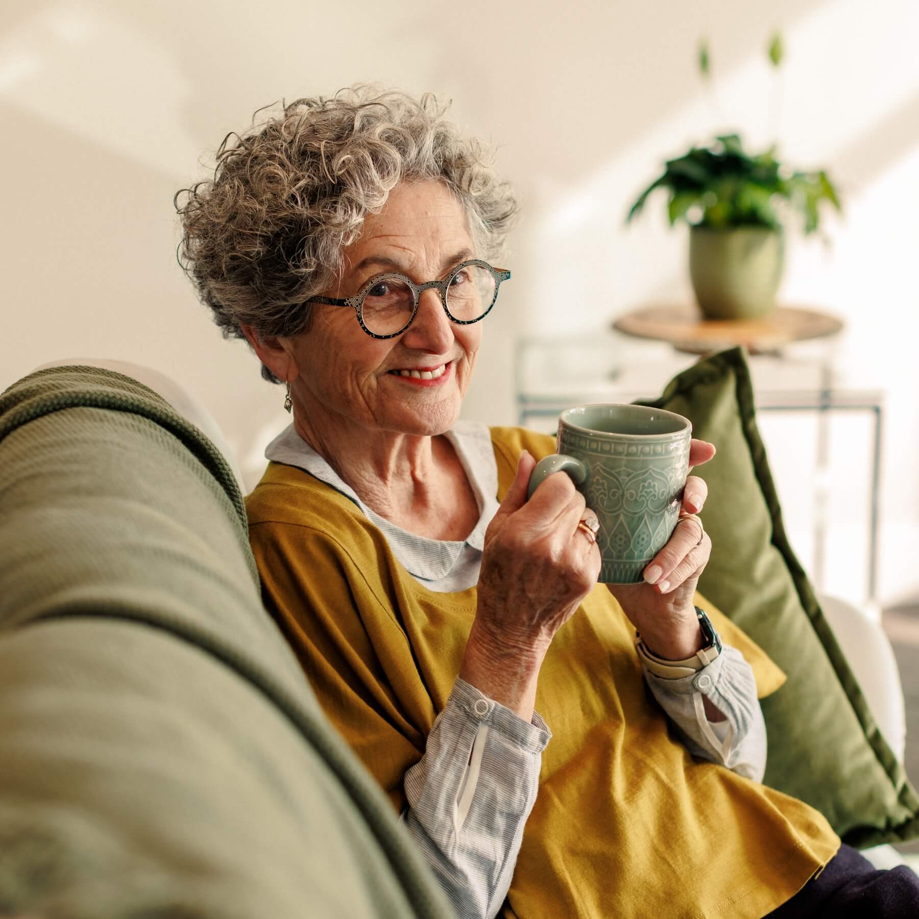 A woman with curly grey hair and glasses smiling at the camera while relaxing on a couch inside her Olive Estate home, holding a mug.