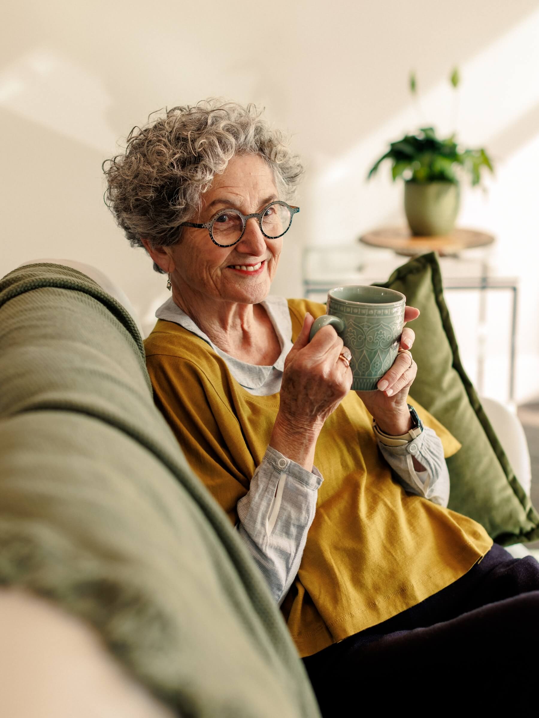 A woman with curly grey hair and glasses smiling at the camera while relaxing on a couch inside her Olive Estate home, holding a mug.