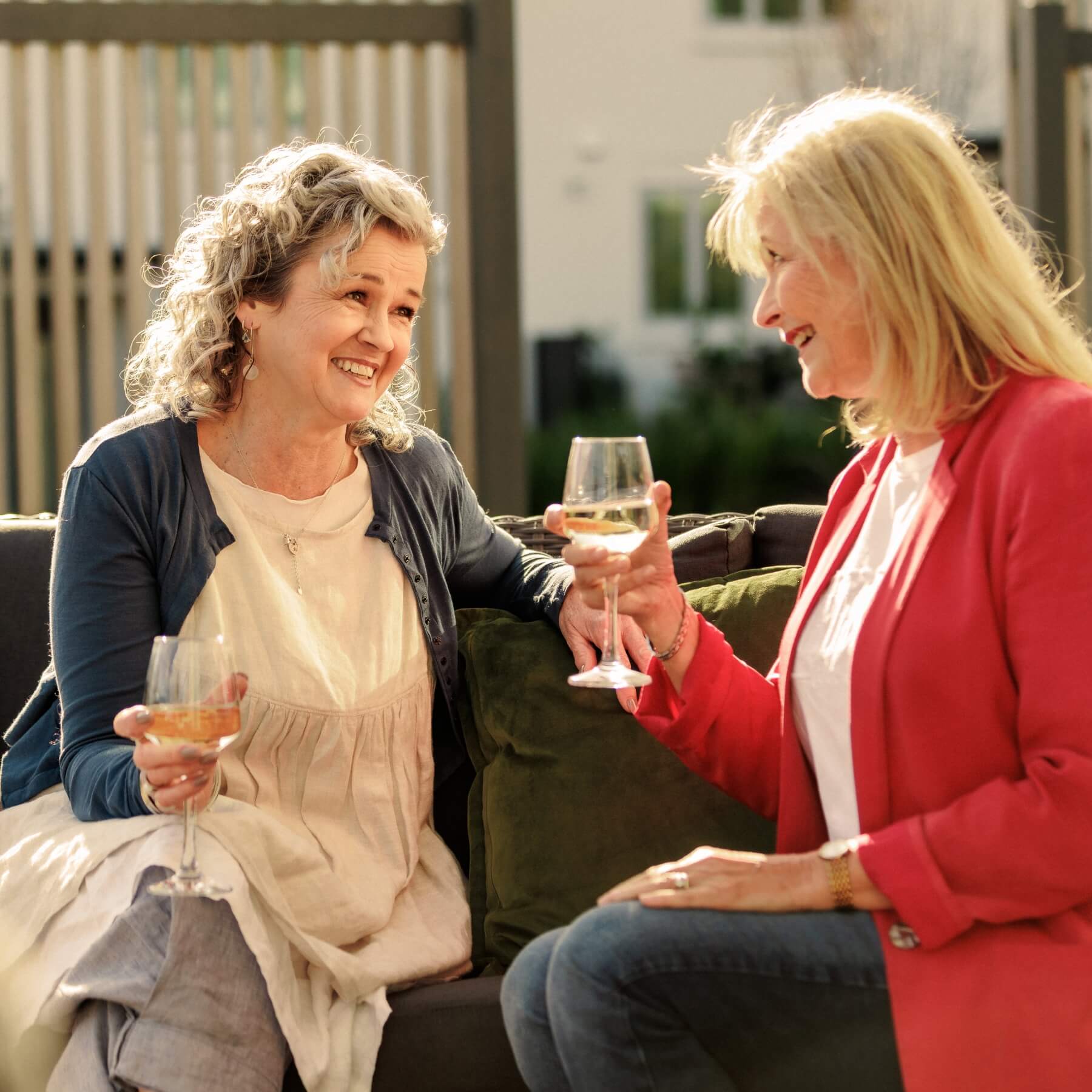 Two women sitting together outdoors on a sunny day, laughing and enjoying glasses of white wine at Olive Estate.