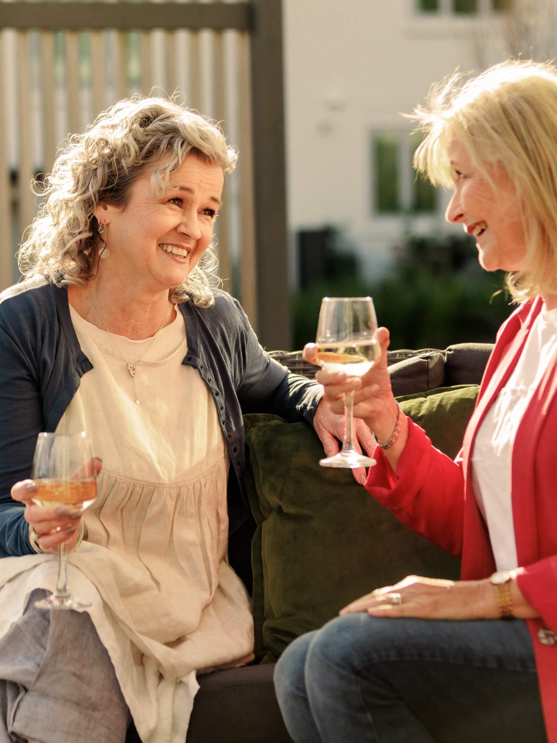 Two women sitting together outdoors on a sunny day, laughing and enjoying glasses of white wine at Olive Estate.