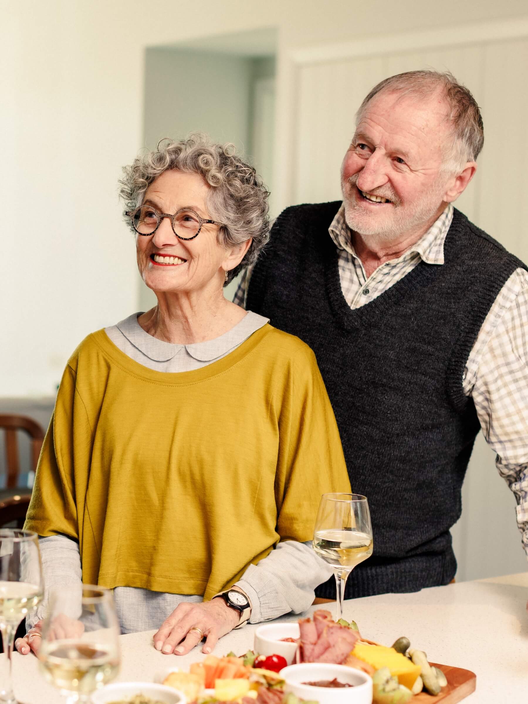 An older couple in a brightly lit Olive Estate kitchen, smiling and socialising around a food spread.