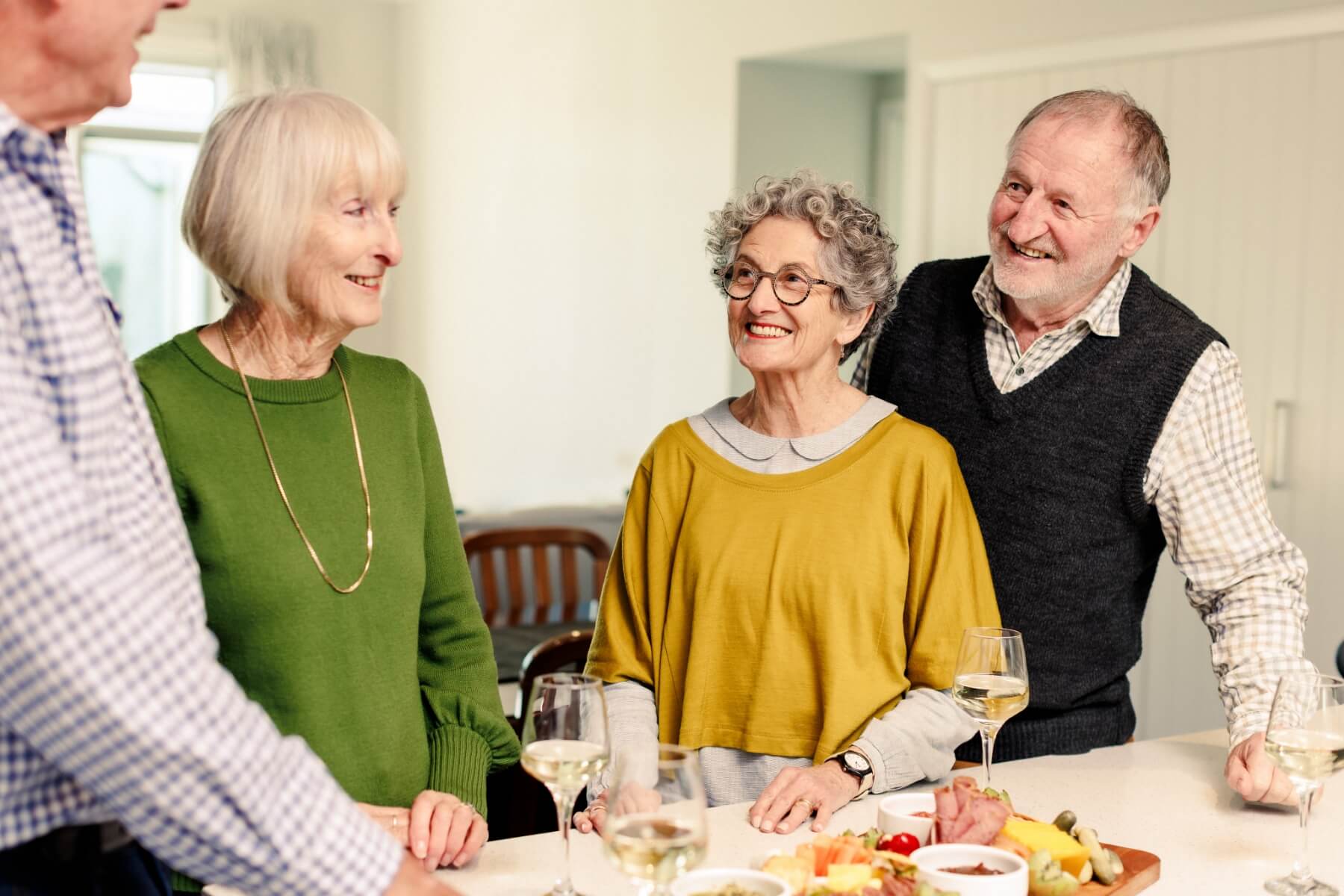 A group of four older adults gathered in a brightly lit Olive Estate kitchen, smiling and socialising around a food spread.